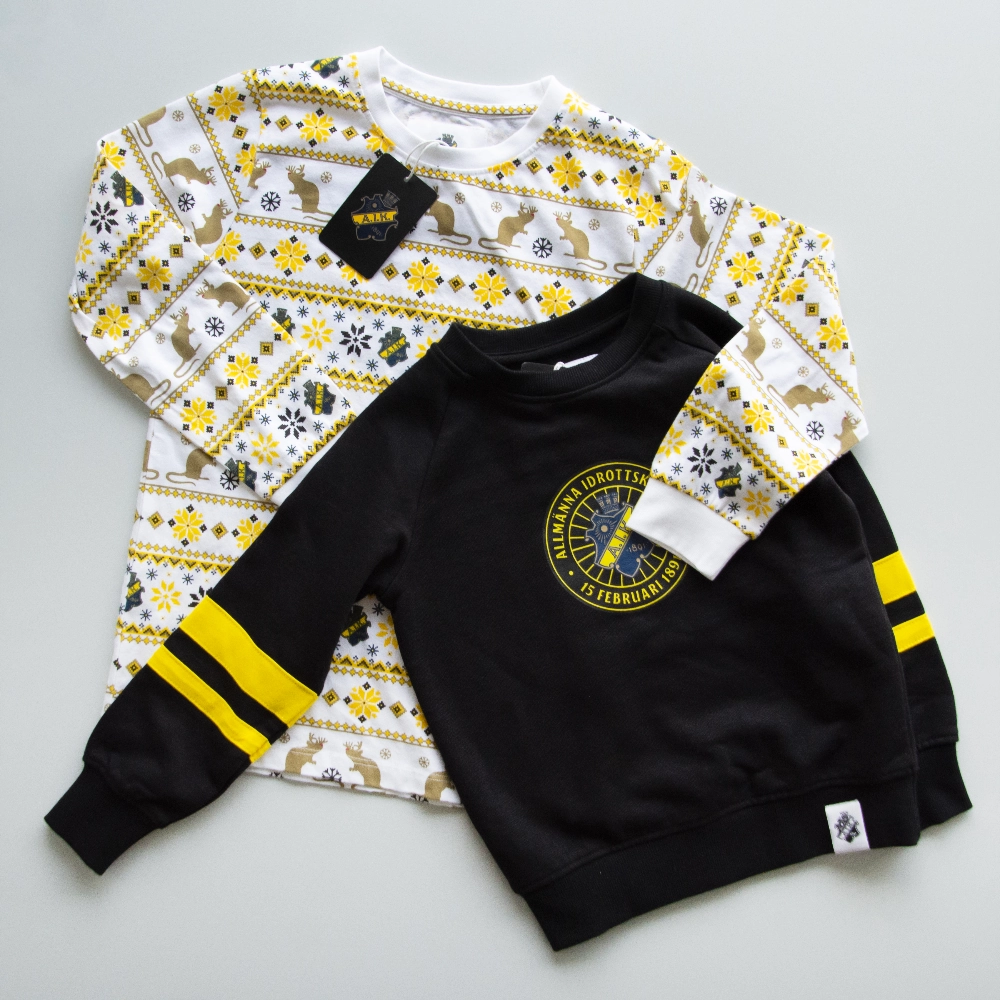  Tv&aring; AIK barntr&ouml;jor p&aring; ett ljust gr&aring;tt underalag: en vit med ren- och sn&ouml;flingsm&ouml;nster i gult och svart, och en svart med gula &auml;rmr&auml;nder och AIK-klubbm&auml;rket p&aring; br&ouml;stet