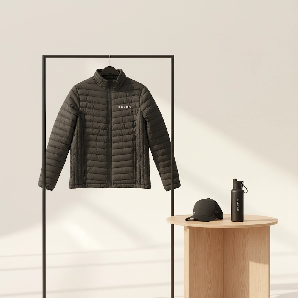 En kviltad Zeekr-jacka p&aring; en kl&auml;dst&auml;llning, med en svart Zeekr-keps och vattenflaska p&aring; ett litet tr&auml;bord.